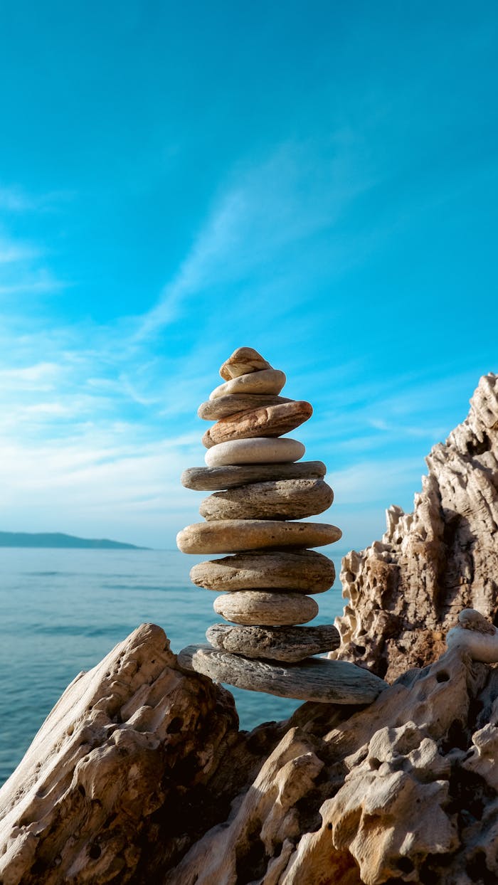 gallery-4 A stack of balanced stones on a rocky seashore with a clear blue sky and sea in the background.